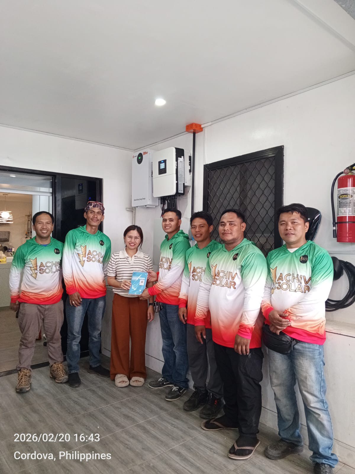 Six Achiv crew lined up with a Cordova homeowner at handover, the customer holding the owner's manual and the finished hybrid inverter mounted on the wall behind them.