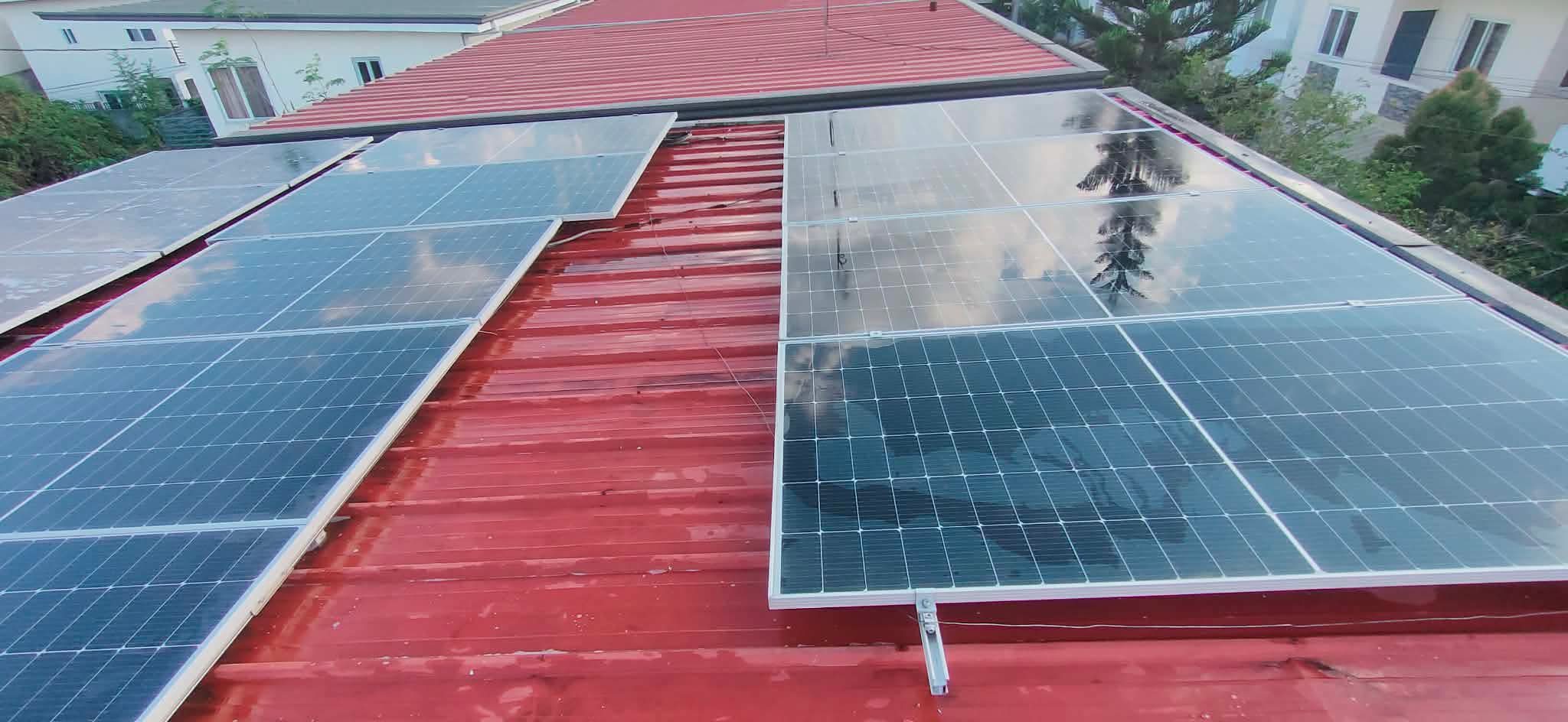 Two clean rows of dark solar panels on a vivid red corrugated roof — a single-family Filipino home with neighbors and trees reflected in the panel glass.