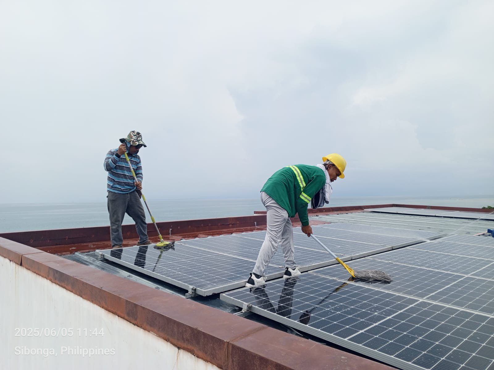 Two cleaners on a flat Sibonga rooftop mopping a row of solar panels, the open ocean horizon stretched behind them under an overcast morning sky.