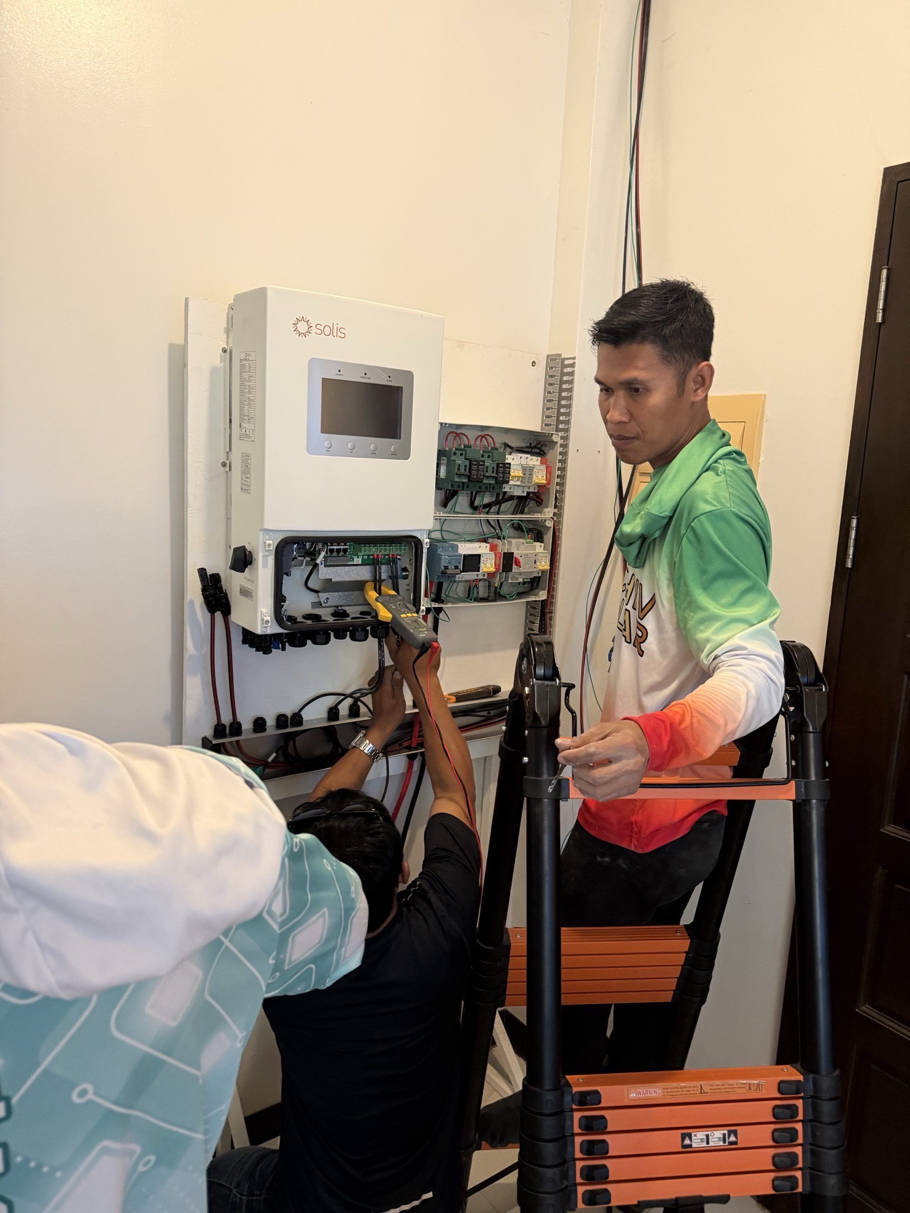 Achiv foreman on a ladder watching a technician below probe an open Solis inverter with a yellow Fluke multimeter — auditing a system left behind by a previous installer.